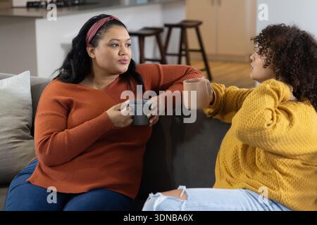 Diverse female friends sitting on gray sofa holding gray and beige mugs, wearing knit sweaters Stock Photo