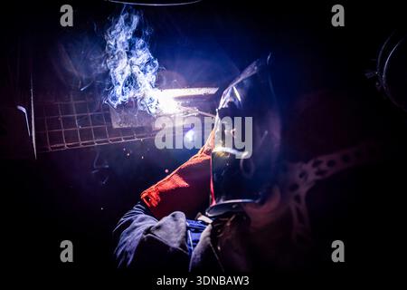 Industrial worker performing arc welding on metal in a dark workshop environment with sparks, smoke and bright welding light. Stock Photo