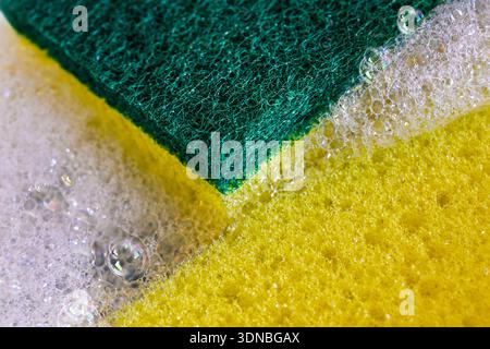 A full-frame, macro photograph focuses on the texture of a yellow and green dual-sided cleaning sponge, surrounded by white soap suds and bubbles, Stock Photo