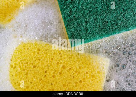 A full-frame, macro photograph focuses on the texture of a yellow and green dual-sided cleaning sponge, surrounded by white soap suds and bubbles, Stock Photo