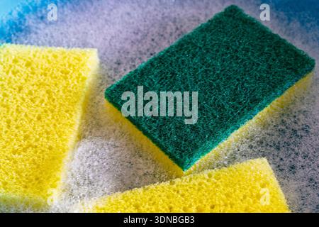 A full-frame, macro photograph focuses on the texture of a yellow and green dual-sided cleaning sponge, surrounded by white soap suds and bubbles, Stock Photo