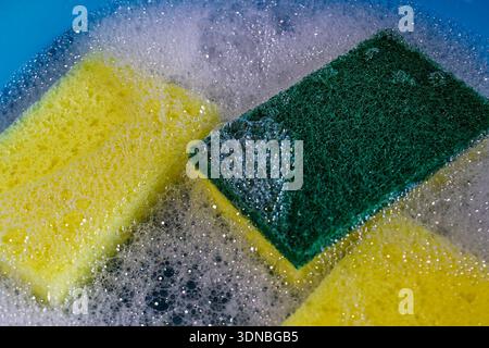 A full-frame, macro photograph focuses on the texture of a yellow and green dual-sided cleaning sponge, surrounded by white soap suds and bubbles Stock Photo