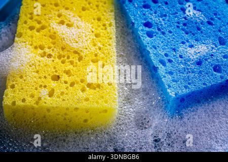 Close-up of a yellow and blue porous cleaning sponge surrounded by thick, foamy white soap bubbles, representing household hygiene, active cleaning. Stock Photo