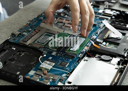 A hand is lifting a metal shield from a laptop motherboard. The workspace shows opened laptop parts and a RAM module is ready to be installed. Stock Photo
