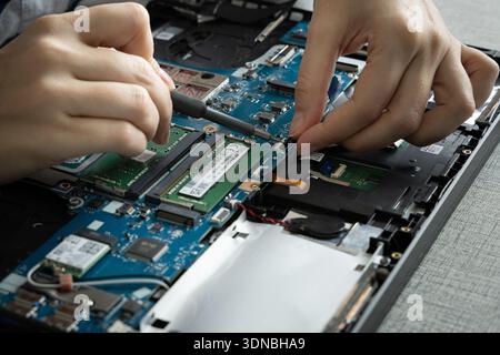 A person works on a laptop at a desk, using a screwdriver to remove components. The hands are focused on replacing the memory and other parts. Stock Photo