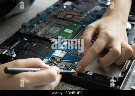 A person is repairing a laptop by inspecting its internal components. The workspace is a flat surface with tools and parts scattered. Stock Photo
