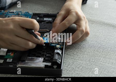 A person is fixing a laptop at a desk. They are using tools to work on the motherboard. Stock Photo