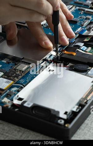 A person uses a screwdriver to open the back of a laptop in a workspace. Various tools and electronic parts are spread out nearby. Stock Photo