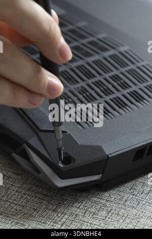 A person uses a screwdriver to remove screws from the back panel of a laptop at home during the afternoon. Stock Photo