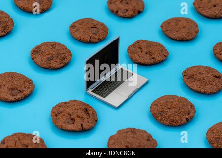 Laptop surrounded by chocolate chip cookies on a blue background. Digital browser cookies, internet privacy, web tracking, and data security concept. Stock Photo