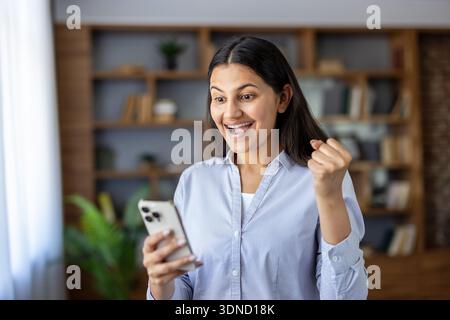 Happy young woman at home reacting to good news on her smartphone, smiling and pumping a fist in celebration of victory, success and joyful achievement Stock Photo