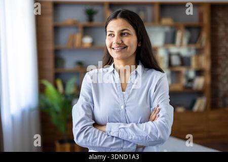 Young professional indian woman standing with arms crossed, smiling confidently and looking away, portraying ambition, success, and positive future vision in a modern office Stock Photo
