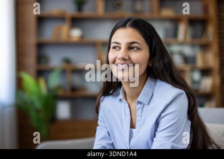 Young indian woman with long dark hair wearing a blue shirt, smiling and looking away while attentively listening in a modern home office setting with bookshelves in the background Stock Photo