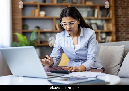 Young indian woman wearing glasses analyzing finances, managing expenses, and preparing tax reports using a laptop and calculator while sitting on a couch in her modern home office Stock Photo