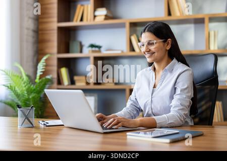 Young businesswoman with dark hair and glasses working on her laptop in a modern wooden office, smiling while typing, showing dedication and efficiency in her professional environment Stock Photo
