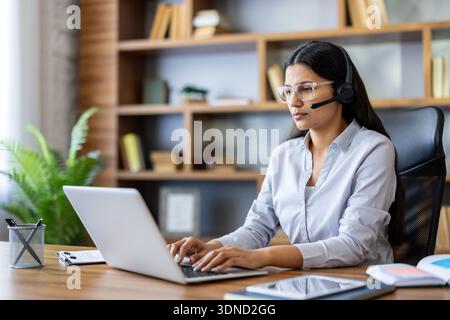 Young indian woman providing customer support or online consultation, wearing a headset with a microphone, using a laptop at a home office desk with a bookshelf in the background Stock Photo