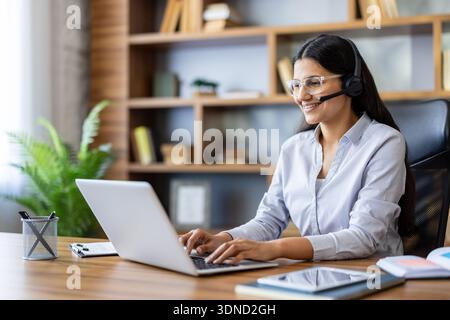 Young woman with headset and glasses cheerfully typing on a laptop, providing customer support and virtual assistance from a modern office desk, professional and friendly Stock Photo