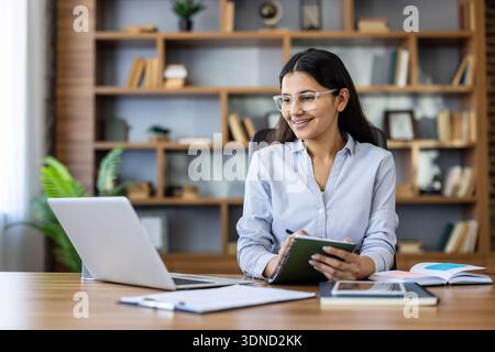 Young professional woman happily working in a modern office, taking notes and looking at her laptop screen, representing productivity, remote work, and business success Stock Photo