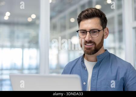 Confident bearded man with glasses smiling while working on a laptop, representing work, focus, technology, and success in a professional office envir Stock Photo