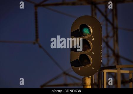 Green railway signal light glowing at dusk with silhouetted industrial structure in the background. Stock Photo