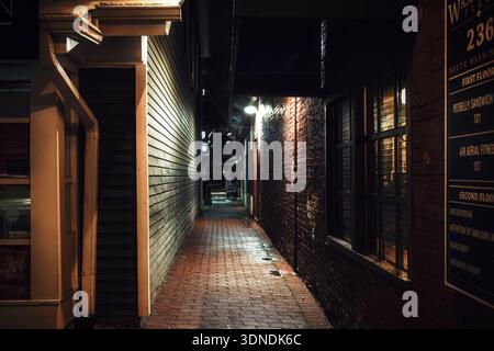Narrow city alley at night illuminated by streetlights, showing brick and wood walls with warm reflected light and deep shadows. Stock Photo
