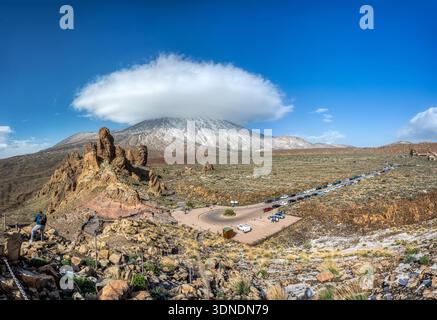 Panoramic view of Roques de García rock formations with snow-covered Teide volcano topped by a distinctive lenticular cloud under a bright blue sky in Stock Photo