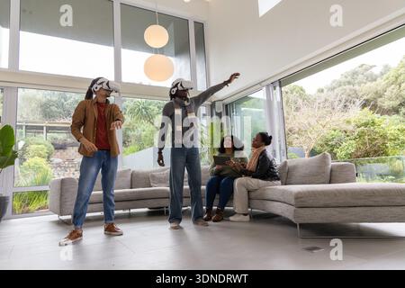 African American family standing using VR headsets while two women sitting on sofa holding tablet Stock Photo