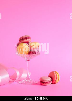 Happy Valentine's day greeting. Pink and yellow color macarons in a champagne glass and pink ribbon on a pink background. Stock Photo