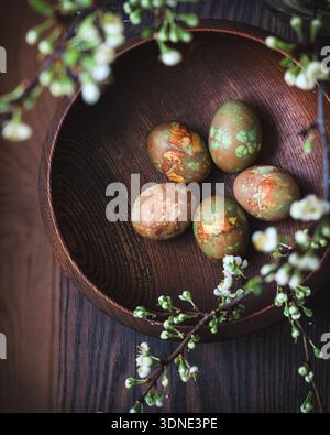 Easter composition with naturally dyed chicken eggs in a dark wooden bowl and blooming plum tree branches in bokeh Stock Photo