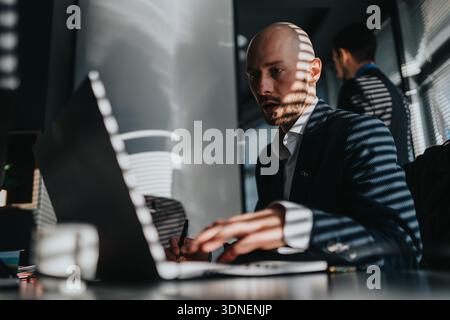 Focused business man works on laptop at a modern office desk with striped light shadows Stock Photo