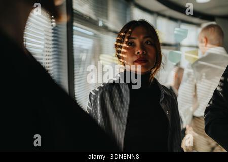 Professional woman in a modern office hallway with blinds, notes, and colleagues Stock Photo