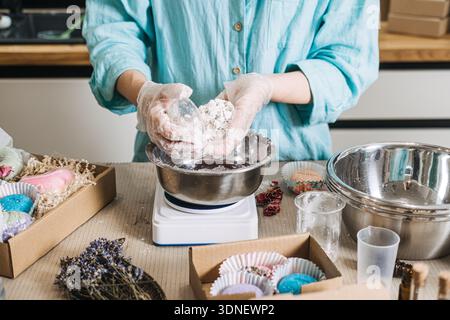 Hands hold large white bath bomb over metal bowl on digital scale surrounded by gift boxes. Artisan formulation, precise handmade, cosmetic craft Stock Photo