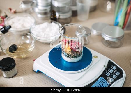 Beaker filled with dried flower petals and white powder sits on digital scale amidst cosmetic supplies. Artisanal flower infusion, slow beauty process Stock Photo