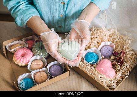 Hands hold heart shaped bath bomb over boxes filled with colorful handmade soaps and shredded paper. personalized beauty gift, diy spa kit, bespoke Stock Photo