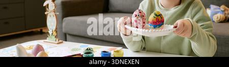 Header of Child Holding Plate with Painted Easter Eggs While Sitting Indoors Stock Photo