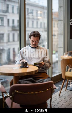 Man book lover fully engaged in reading popular book while sitting in cozy cafe with cup of coffee. Stock Photo