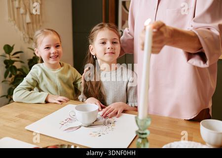 Two Girls Smiling While Sitting at Table with Adult Lighting Candle Stock Photo