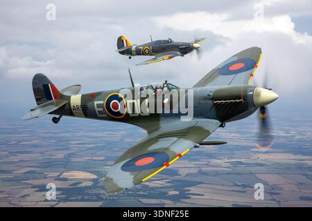 The Supermarine Spitfire LF5C, G-AWII, and Hawker Sea Hurricane Mk1B, G-BKTH, fly in formation at UK Warbirds 2023. The Spitfire, with Rolls-Royce Merlin 66 engine and elliptical wings, represents RAF fighter design, while the Sea Hurricane, powered by Merlin 32, reflects Royal Navy carrier-based aviation. The air-to-air close-up highlights pilot cockpits, fuselage structures, and wing profiles, showcasing historical WWII aircraft and formation flight operations. Stock Photo
