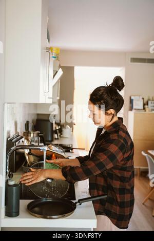 Woman wearing a plaid shirt washes a glass lid in a modern kitchen, focusing on household chores. The kitchen setup includes various utensils and a cl Stock Photo