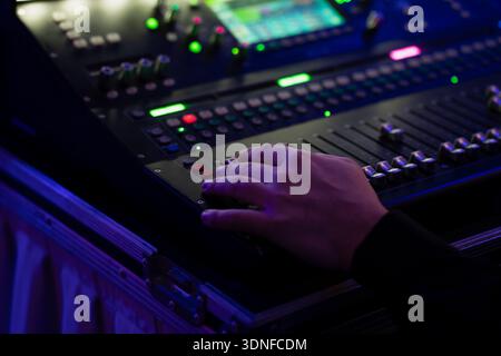 Hand operating professional audio mixing console with faders and glowing buttons in flight case, close up live event sound control under blue light Stock Photo
