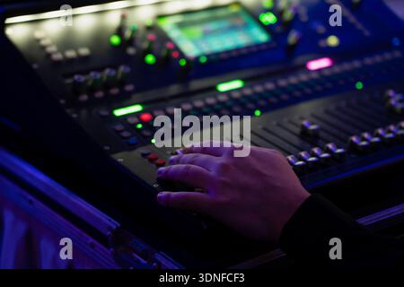Hand on digital audio mixing console in road case, adjusting faders and backlit buttons for live event sound control under blue stage lighting Stock Photo