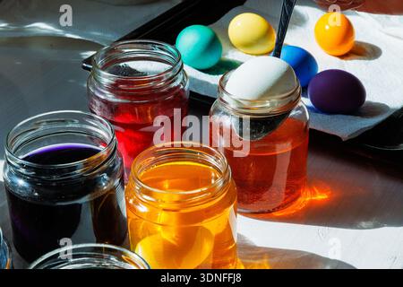 Spoon putting white egg into an orange dye jar with colorful dyed easter eggs nearby Stock Photo
