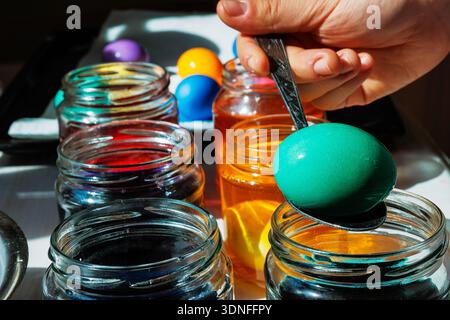 Close up of a hand lifting a green dyed easter egg on a spoon above a glass jar, with orange dye jars glowing in sunlight behind. Stock Photo