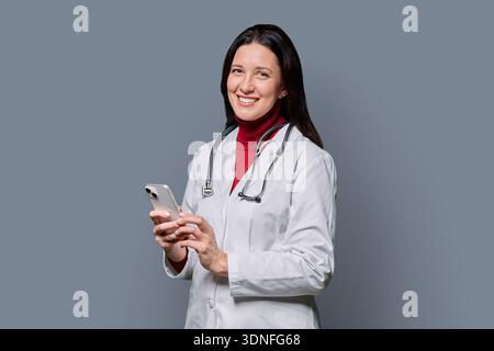 Middle aged female doctor holding smartphone, on grey studio background Stock Photo