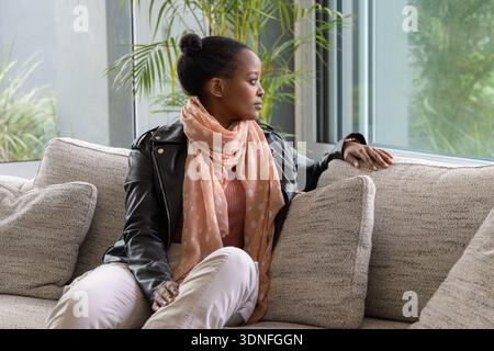 African American woman sitting on beige sofa looking out large window wearing leather jacket, scarf Stock Photo