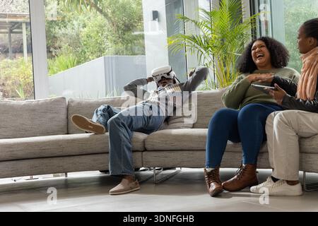 African American friends laughing, reclining on grey sofa in living room, one using VR headset Stock Photo