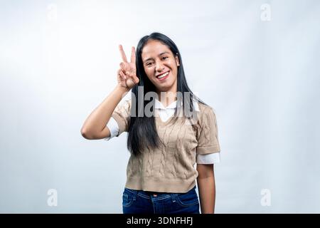 Happy young Southeast Asian woman smiling at camera and showing victory or peace sign gesture against a clean white background. Stock Photo