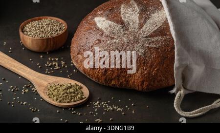Fresh baked round loaf of spelt bread with crushed hemp seeds.Bread decorated with cannabis leaf made of flour in hemp cloth bag and spoon with pulver Stock Photo