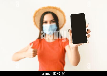 Young woman wearing protective face mask and hat, showing smartphone with empty blank screen on light wall Stock Photo