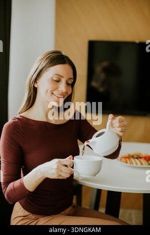 A woman sits at a table and pours tea into a cup. She smiles while looking at the cup. Snacks are placed on the table beside her in a room with warm l Stock Photo